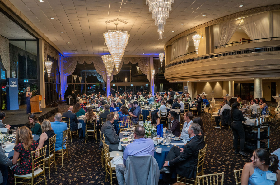 A speaker presents to a large audience seated at round tables in an elegant ballroom with chandeliers.